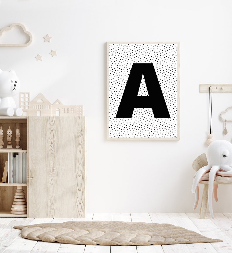 May include: A framed print featuring a large black letter "A" against a white background speckled with small black triangles. The print hangs on a white wall in a child's room, alongside wooden toys and a rug.