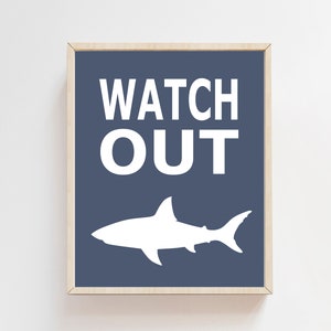 Op de afbeelding: Een ingelijst print met een blauwe achtergrond en witte tekst die "WATCH OUT" luidt. Onder de tekst bevindt zich een wit silhouet van een haai.