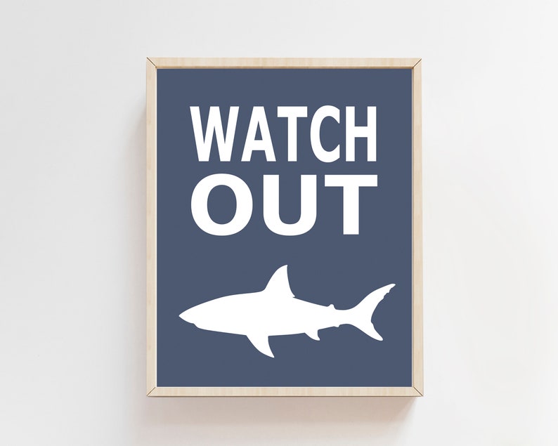 Op de afbeelding: Een ingelijst print met een blauwe achtergrond en witte tekst die "WATCH OUT" luidt. Onder de tekst bevindt zich een wit silhouet van een haai.