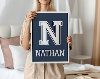 May include: A framed navy blue print with a white border. The print features a large white letter "N" above the name "NATHAN" in white, all in a collegiate style font. The frame is held by a person.