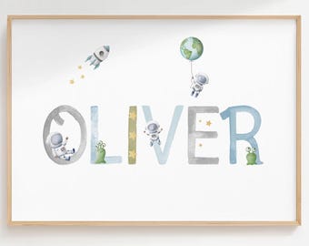 May include: Watercolor print featuring the name "OLIVER" in large, whimsical letters. The letters are decorated with space-themed illustrations, including astronauts, a rocket, stars, and a planet. The artwork is framed in a light wood.