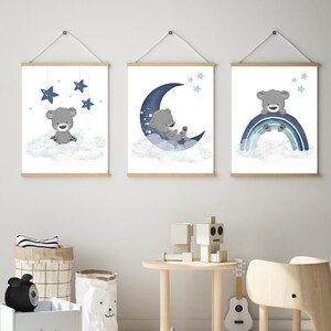 Op de afbeelding: Drie ingelijste prints met een blauw en grijs kleurenschema met een cartoonbeer op een wolk, een slapende beer op een halve maan en een beer die op een regenboog zit. De prints hangen aan een witte muur.