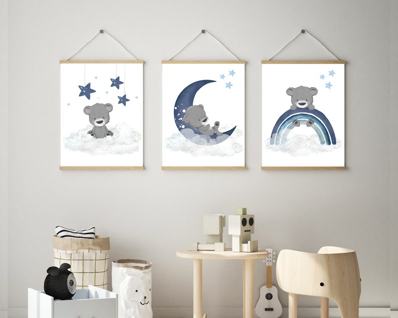 Op de afbeelding: Drie ingelijste prints met een blauw en grijs kleurenschema met een cartoonbeer op een wolk, een slapende beer op een halve maan en een beer die op een regenboog zit. De prints hangen aan een witte muur.