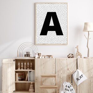 May include: A framed print featuring a large black letter "A" against a background of small black triangles. The print hangs above a wooden cabinet filled with toys, including a rainbow abacus and wooden train. A small wooden step stool sits in front.