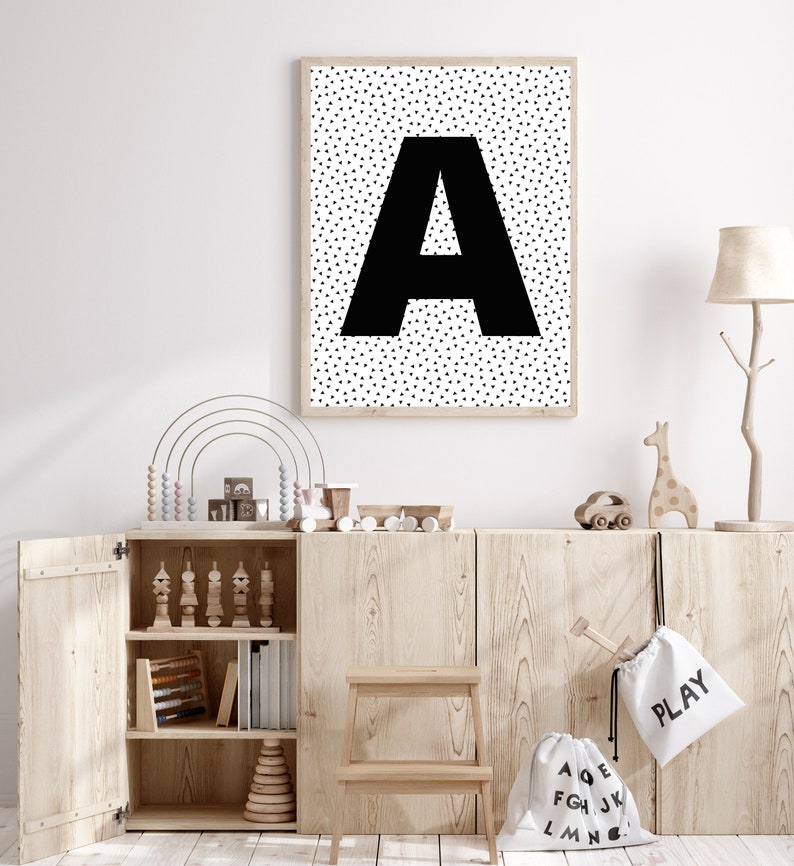 May include: A framed print featuring a large black letter "A" against a background of small black triangles. The print hangs above a wooden cabinet filled with toys, including a rainbow abacus and wooden train. A small wooden step stool sits in front.