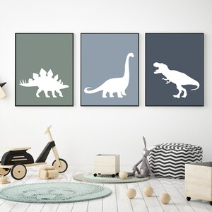 May include: Three framed prints of white dinosaur silhouettes on different colored backgrounds. The prints are hung on a white wall in a child's room. The room also features a wooden toy scooter, a round rug, and a beanbag chair.