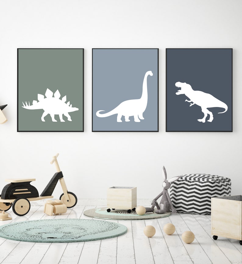 May include: Three framed prints of white dinosaur silhouettes on different colored backgrounds. The prints are hung on a white wall in a child's room. The room also features a wooden toy scooter, a round rug, and a beanbag chair.