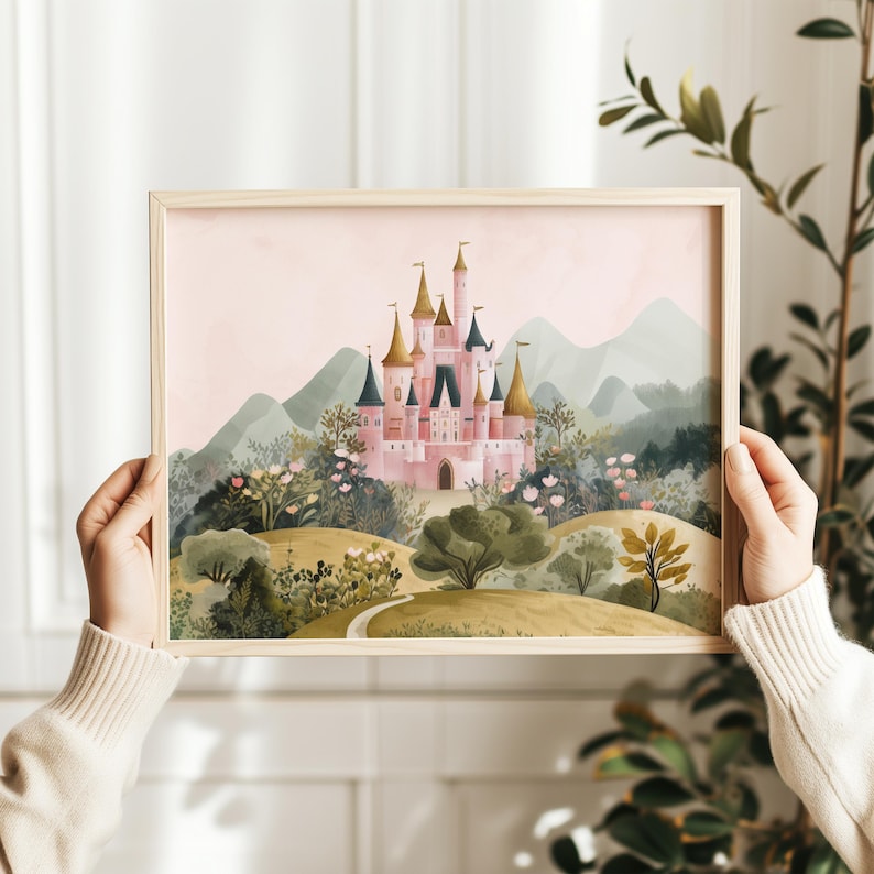 May include: A framed art print featuring a pink castle with gold-tipped towers, set amidst a lush landscape of green trees, flowers, and rolling hills. The background shows soft, muted mountains under a pale pink sky. The frame is made of light-colored wood.
