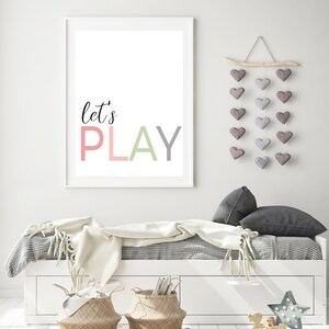 May include: A white framed print with the text "let's PLAY" in pink, green, and gray letters. The print is hanging on a white wall above a white daybed with a gray and white striped bedspread and a white blanket. There is a wooden branch with gray felt hearts hanging from it on the wall.