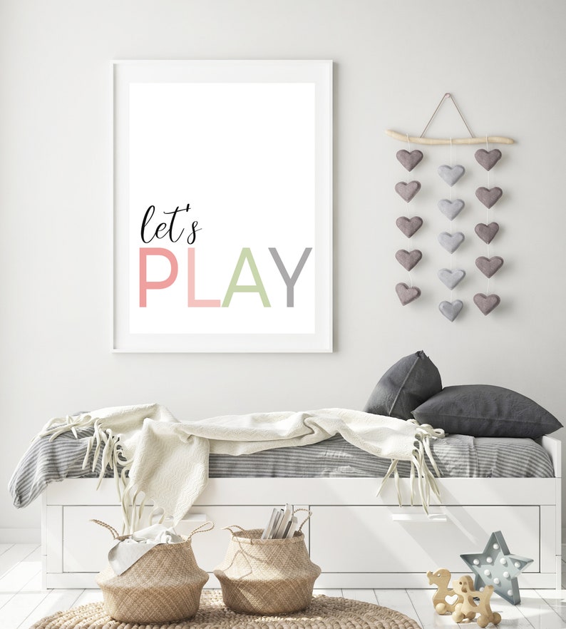 May include: A white framed print with the text "let's PLAY" in pink, green, and gray letters. The print is hanging on a white wall above a white daybed with a gray and white striped bedspread and a white blanket. There is a wooden branch with gray felt hearts hanging from it on the wall.