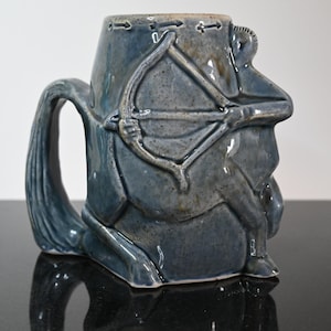May include: A blue ceramic mug featuring a sculpted design of a person riding a horse and holding a bow and arrow. The mug has a handle shaped like a horse's tail.