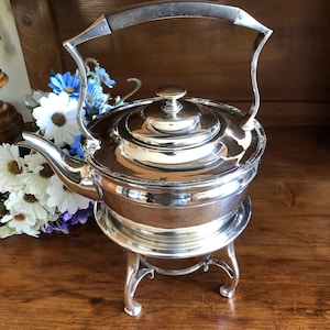 May include: A silver teapot with a wooden handle and a stand. The teapot is on a wooden surface with a bouquet of white and blue flowers.