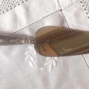 Puede incluir: Servidor de pastel plateado con un mango decorativo y una hoja ancha y plana. El mango presenta diseños florales y de volutas. El servidor está sobre un mantel blanco bordado.