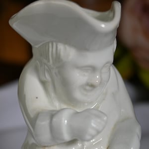 Könnte beinhalten: Weiße Porzellanfigur einer Person mit Hut. Die Person lächelt und hat die Arme verschränkt.