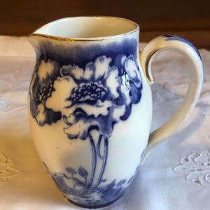 May include: A white ceramic milk jug with a blue and white floral design. The jug has a gold rim and a handle.