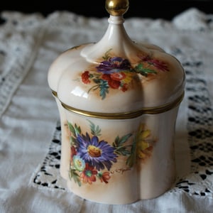 Puede incluir: Un tarro de cerámica blanca con tapa dorada y estampado floral. El tarro tiene un borde festoneado y está decorado con una variedad de flores en tonos de rosa, morado, amarillo y rojo.
