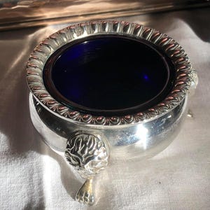 Op de afbeelding: Een zilveren zoutkelder met een diepblauwe glazen binnenkant. De rand is versierd met een touwachtig ontwerp. De zijkanten hebben sierlijke leeuwenkopdetails met kleine voetjes. Het stuk is waarschijnlijk antiek.