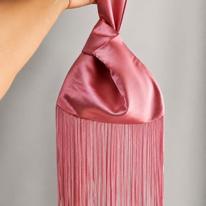 May include: A dusty rose satin knot bag featuring a smooth, lustrous finish and a long, flowing fringe trim along the bottom edge. The bag is constructed with a single handle loop that creates a gathered, knotted effect at the top opening.