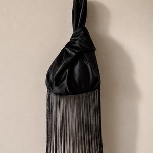 May include: A black satin handbag featuring a knotted handle design and long, thin black fringe detailing that hangs from the base of the bag. The fabric has a smooth, reflective finish and is suspended from a gold-toned metal hook.