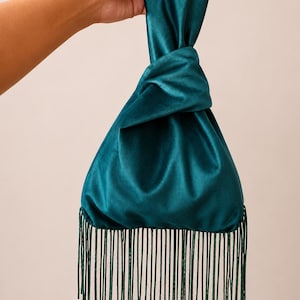 May include: A teal velvet handbag featuring a knotted top handle design and a long, straight fringe trim along the bottom edge. The fabric has a soft, reflective sheen, and the fringe consists of numerous thin, matching teal threads.