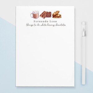 Peut inclure: Un bloc-notes blanc avec le texte "Fernando Leon things to do while having chocolate" et des illustrations de chocolat chaud, de barres chocolatées et de biscuits. Un stylo blanc est à droite du bloc-notes. Le fond est bleu clair et blanc.