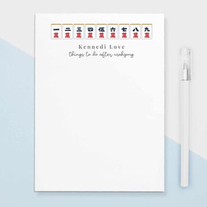 May include: A white notepad with a blue and red mahjong tile design at the top. The notepad has the text "Kennedi Love" and "things to do after mahjong" written on it. A white pen is lying next to the notepad.