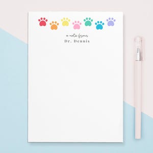 May include: A white notepad with a rainbow paw print border and the text "a note from Dr. Dennis". The notepad is on a light blue and pink background with a white pen.