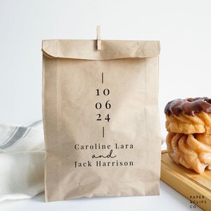 May include: A brown paper bag with a white label that says "10 06 24" and "Caroline Lara and Jack Harrison". The bag is 12.7 cm by 20.3 cm and is shown in a photo.