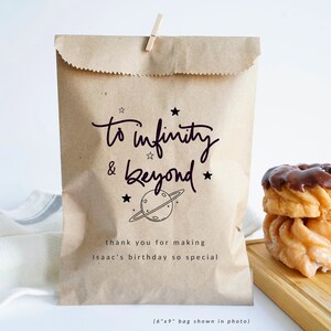 May include: A brown paper gift bag with a black and white illustration of a planet and stars. The bag has the text "to infinity & beyond" and "thank you for making Isaac's birthday so special". The bag is 15.2 cm by 22.9 cm.