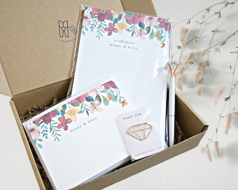 May include: A brown cardboard box with a floral patterned notepad, a pen, and a gold paper clip. The notepad has the text "a note from Diane M Kaye" printed on it. The box is filled with brown packing paper.