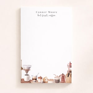 May include: A rectangular notepad with a white background featuring the printed text "Conner Moore" and "but first, coffee" at the top. The bottom border is decorated with a watercolour illustration of various coffee brewing equipment and cups.
