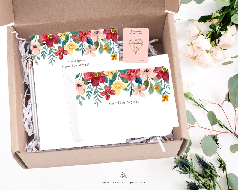 May include: A gift box filled with white paper shred, a pen, a set of floral stationery with the name "Camille Wyatt" printed on the cards, and a diamond-shaped paper clip. The box is open and the items are visible.