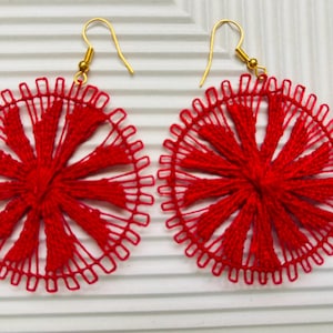 May include: Pair of red, circular earrings with a woven design. The earrings have a sunburst pattern and gold-colored hooks. The earrings are displayed on a white, textured surface. These earrings are a unique accessory.