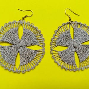 May include: A pair of handmade gray earrings with a circular design. Each earring features a woven, four-petal flower shape at the center, surrounded by a detailed, scalloped edge. The earrings have gold-colored hooks and are set against a bright yellow background.