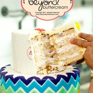 Puede incluir: Un pastel de dos pisos con una base de fondant blanco y un patr&oacute;n de chevron arco&iacute;ris en el piso inferior. El piso superior est&aacute; parcialmente cortado, revelando capas de pastel y glaseado. El pastel est&aacute; decorado con un peque&ntilde;o coraz&oacute;n rojo y el texto "Beyond Buttercream".