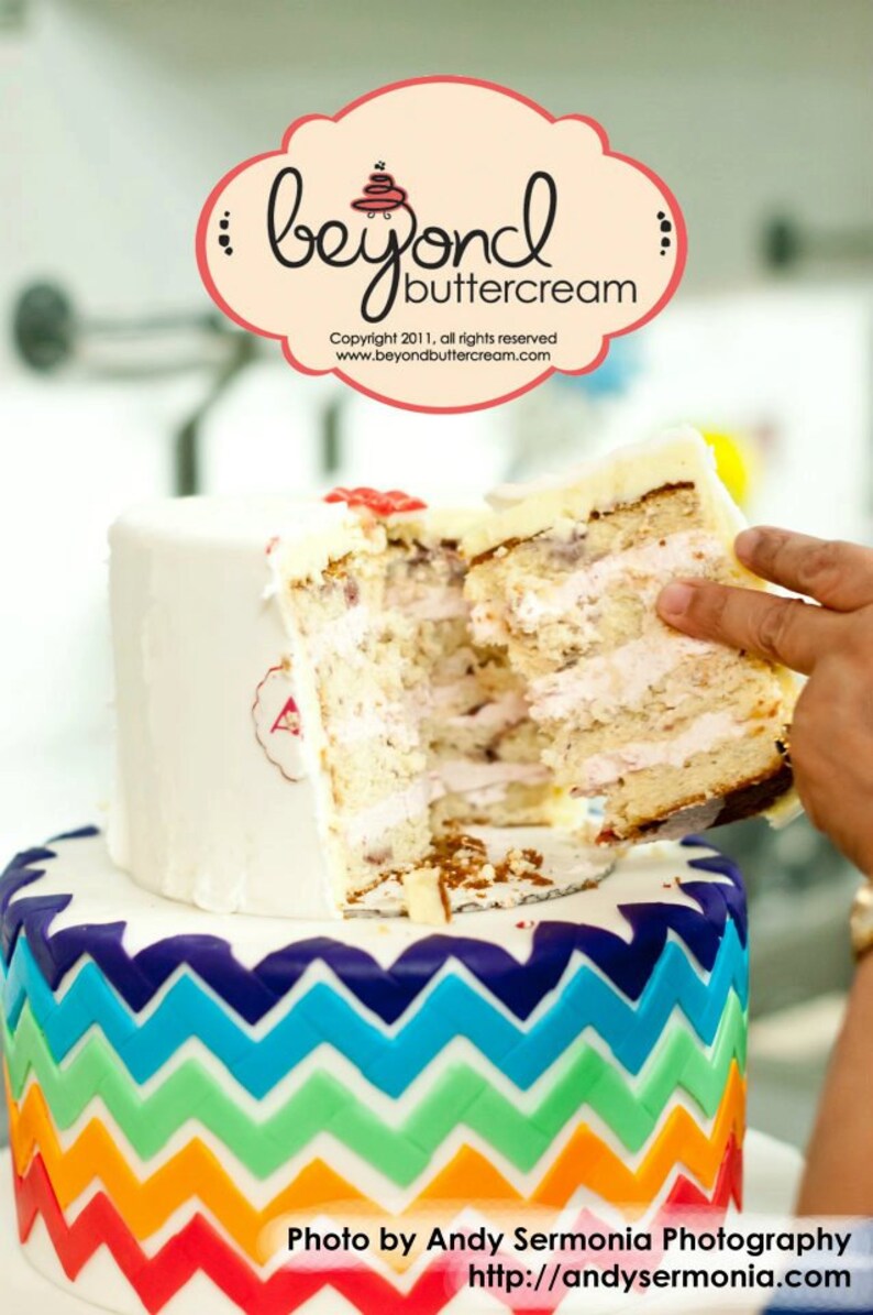 Puede incluir: Un pastel de dos pisos con una base de fondant blanco y un patr&oacute;n de chevron arco&iacute;ris en el piso inferior. El piso superior est&aacute; parcialmente cortado, revelando capas de pastel y glaseado. El pastel est&aacute; decorado con un peque&ntilde;o coraz&oacute;n rojo y el texto "Beyond Buttercream".