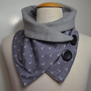 Puede incluir: Una bufanda gris y blanca de cuello de tortuga con un patrón de anclas blancas y dos botones negros. La bufanda está hecha de un tejido suave forrado de polar.