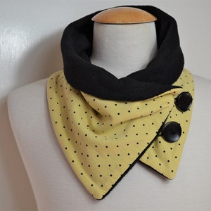 Puede incluir: Bufanda amarilla y negra con estampado de lunares y dos grandes botones negros. La bufanda está envuelta alrededor del cuello de un maniquí, mostrando su diseño y estilo. Hecha de tela suave.