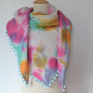 Può includere: Una sciarpa triangolare tie-dye con sfondo bianco e motivi tie-dye rosa, blu, verde e giallo. La sciarpa ha un bordo di pon pon blu chiaro.