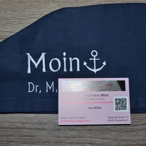 Könnte beinhalten: Eine rosafarbene Visitenkarte mit dem Text "Sand-mère Mimi" und einem QR-Code. Der Text "Mobil: +491777313855" und "grandmeremimi@gmx.de" befinden sich ebenfalls auf der Karte. Die Karte liegt auf einem dunkelblauen Stoff mit dem gestickten Text "Moin" und einem Anker.