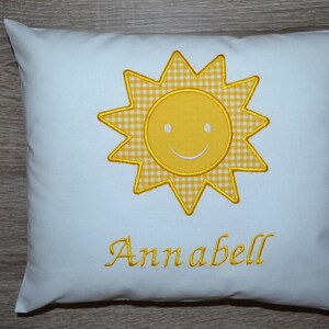 Puede incluir: Almohada blanca con un sol bordado amarillo con una cara sonriente y el nombre Annabell.