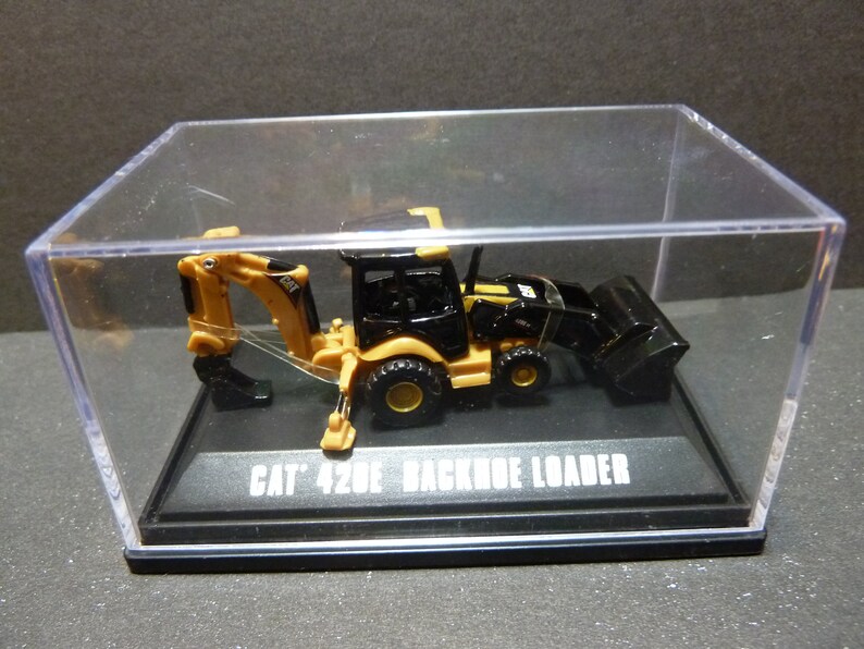 May include: A miniature CAT 420E backhoe loader model, predominantly black and yellow, encased in a clear display case. The model sits on a black base with the text "CAT 420E BACKHOE LOADER". The backhoe arm is extended.