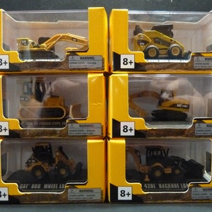 May include: Six boxed toy construction vehicles, including an excavator, bulldozer, and backhoe loader. Each toy is yellow and black, with the CAT logo visible. The boxes are yellow with clear plastic windows and the text "8+" and a choking hazard warning.