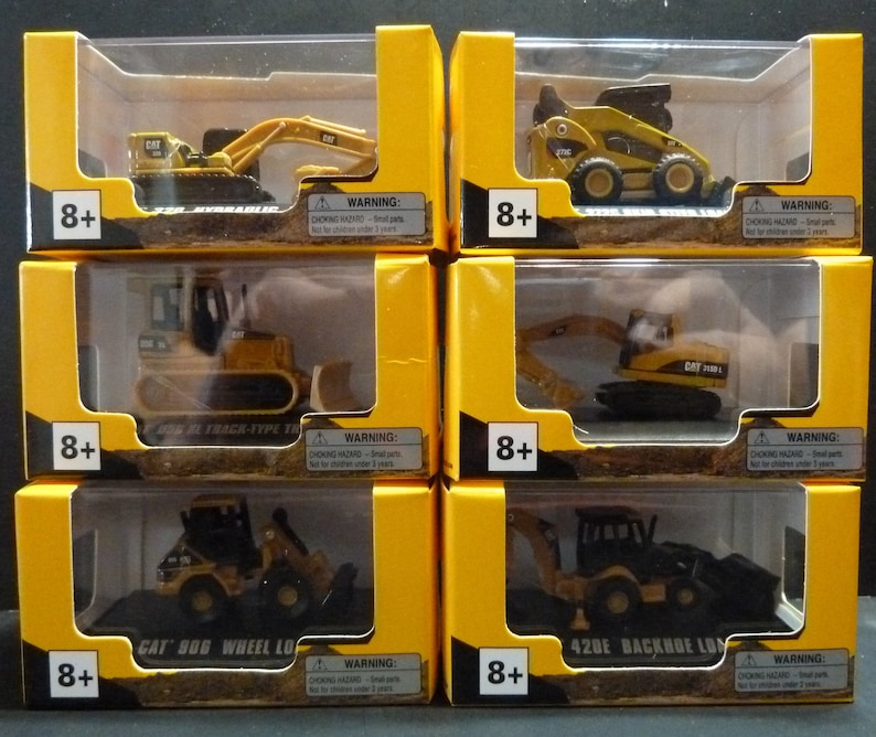 May include: Six boxed toy construction vehicles, including an excavator, bulldozer, and backhoe loader. Each toy is yellow and black, with the CAT logo visible. The boxes are yellow with clear plastic windows and the text "8+" and a choking hazard warning.
