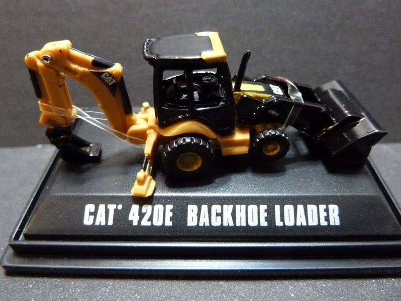 May include: A miniature CAT 420E Backhoe Loader model. The construction vehicle is black and yellow, with a detailed design. The model sits on a black base with the text "CAT 420E BACKHOE LOADER" in white.
