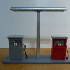 May include: A model of a gas station with two pumps, one gray and one red, with a silver canopy overhead. The pumps have digital displays and hoses.