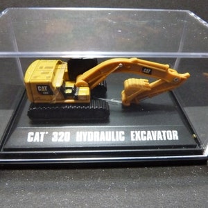 May include: A yellow CAT 320 hydraulic excavator model displayed in a clear acrylic case. The model has black tracks and a movable arm. The text "CAT 320 HYDRAULIC EXCAVATOR" is printed on the base.