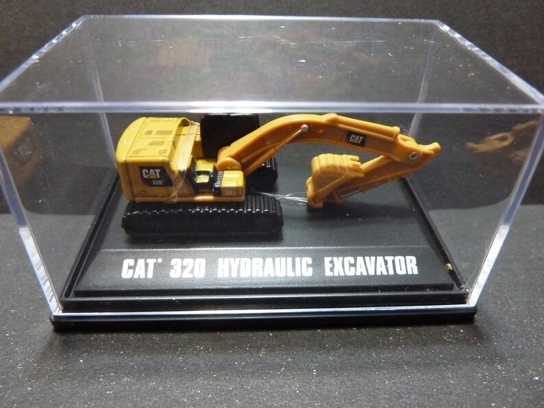 May include: A yellow CAT 320 hydraulic excavator model displayed in a clear acrylic case. The model has black tracks and a movable arm. The text "CAT 320 HYDRAULIC EXCAVATOR" is printed on the base.