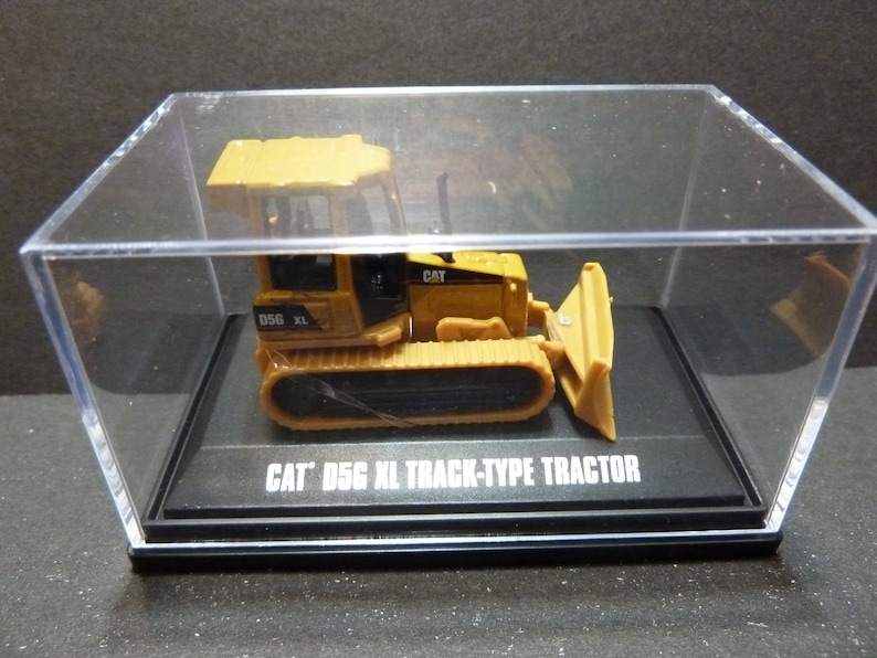 May include: A yellow and black CAT D5G XL track-type tractor model is displayed in a clear acrylic case. The model features a blade and track design, with the text "CAT D5G XL TRACK-TYPE TRACTOR" on the base.