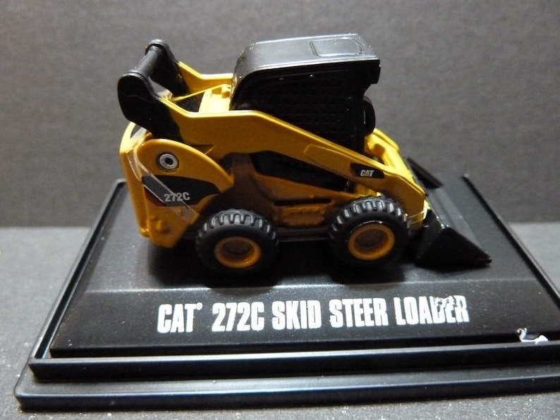 May include: A yellow and black CAT 272C skid steer loader model. The model has black tires, a black cab, and a black bucket. The text "CAT 272C SKID STEER LOADER" is printed in white on the black base.